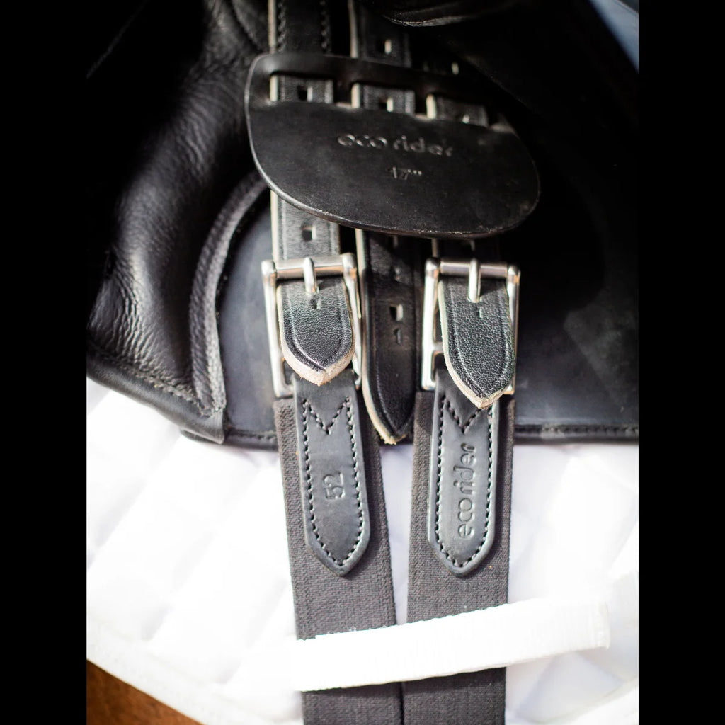 Close-up of the black EcoRider girth buckled to the saddle billets, showing the size 52 stamp and debossed branding.